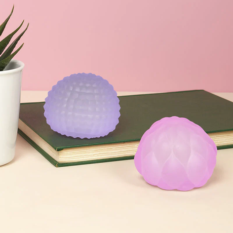 Purple and pink silicone stress balls with textured surfaces sit on a green book. A small plant is on the left, against a pink background.