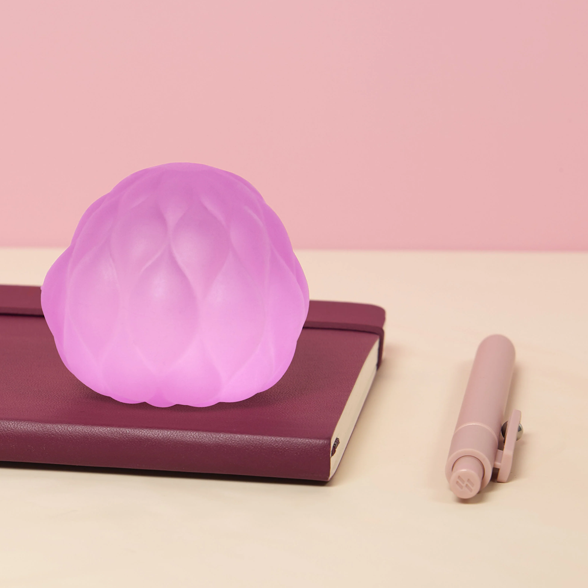 A soft, glowing pink orb rests on a closed burgundy notebook on a beige surface. A light pink pen lies beside it. The background is a pale pink, creating a calming tone.