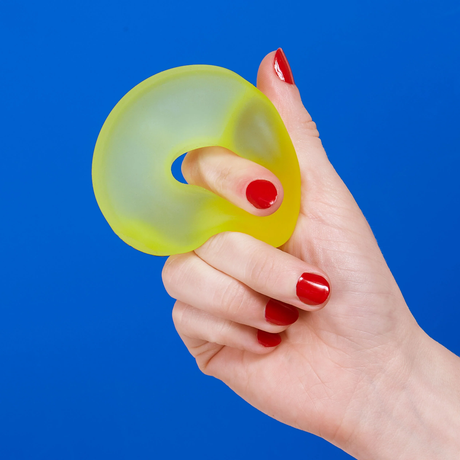 Hand squeezing a yellow Speks Loop Gump memory gel stress ring toy on a blue background.