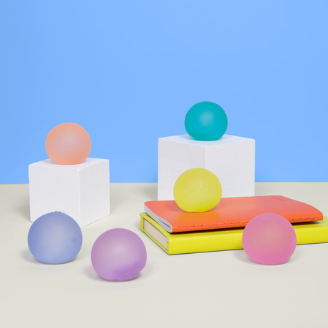 Colorful pastel balls are displayed on white cubes and stacked notebooks against a blue background. The scene is playful and artistic.