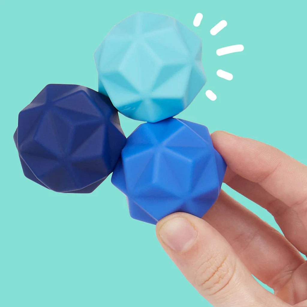 A hand holds three geometric stress balls in dark blue, light blue, and teal against a teal background. The mood is calming and playful.