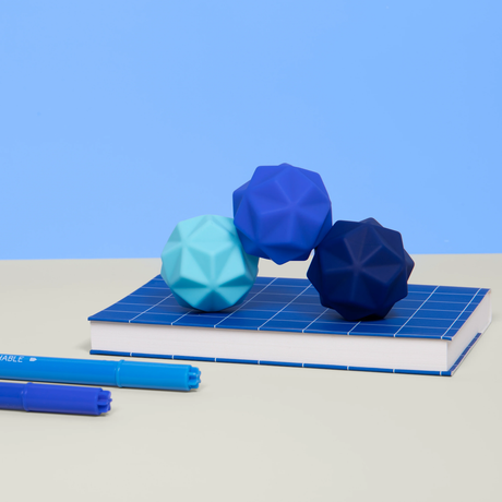 Geometric blue stress balls on a blue-checkered notebook, bathed in soft lighting against a blue backdrop. Two blue pens lie parallel in the foreground, creating a calming atmosphere.