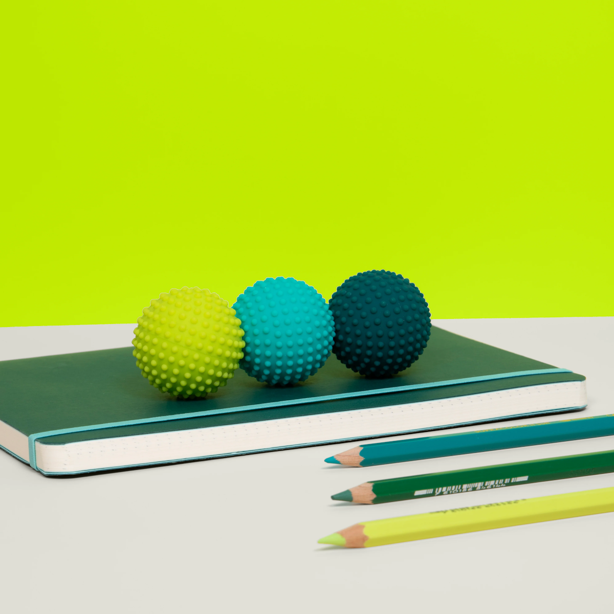 Three textured balls in yellow, teal, and dark blue sit on a green notebook, with matching colored pencils nearby. Bright lime background creates a playful vibe.
