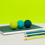 Three textured balls in yellow, teal, and dark blue sit on a green notebook, with matching colored pencils nearby. Bright lime background creates a playful vibe.