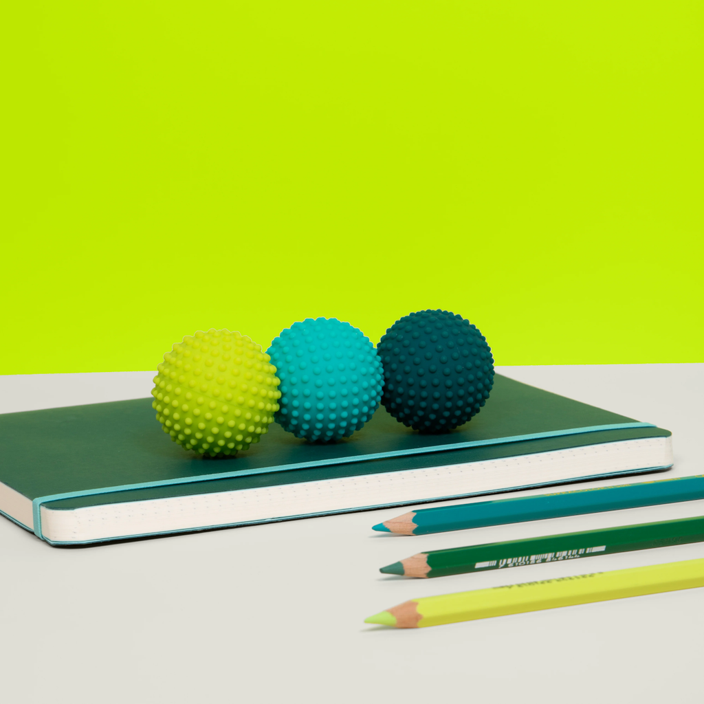 Three textured balls in yellow, teal, and dark blue sit on a green notebook, with matching colored pencils nearby. Bright lime background creates a playful vibe.