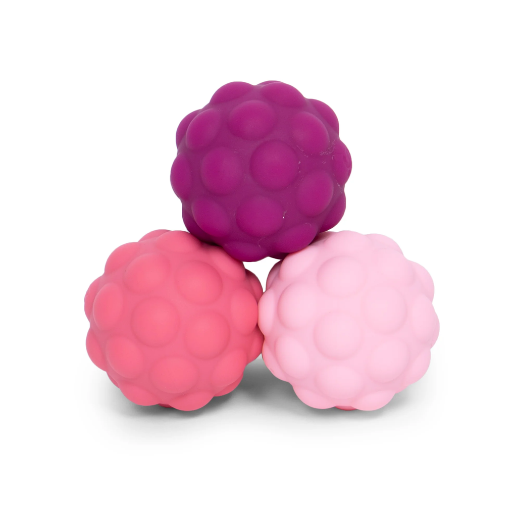 Three textured massage balls stacked in a pyramid. Colors are deep purple, bright pink, and light pink. The tone is playful and tactile.