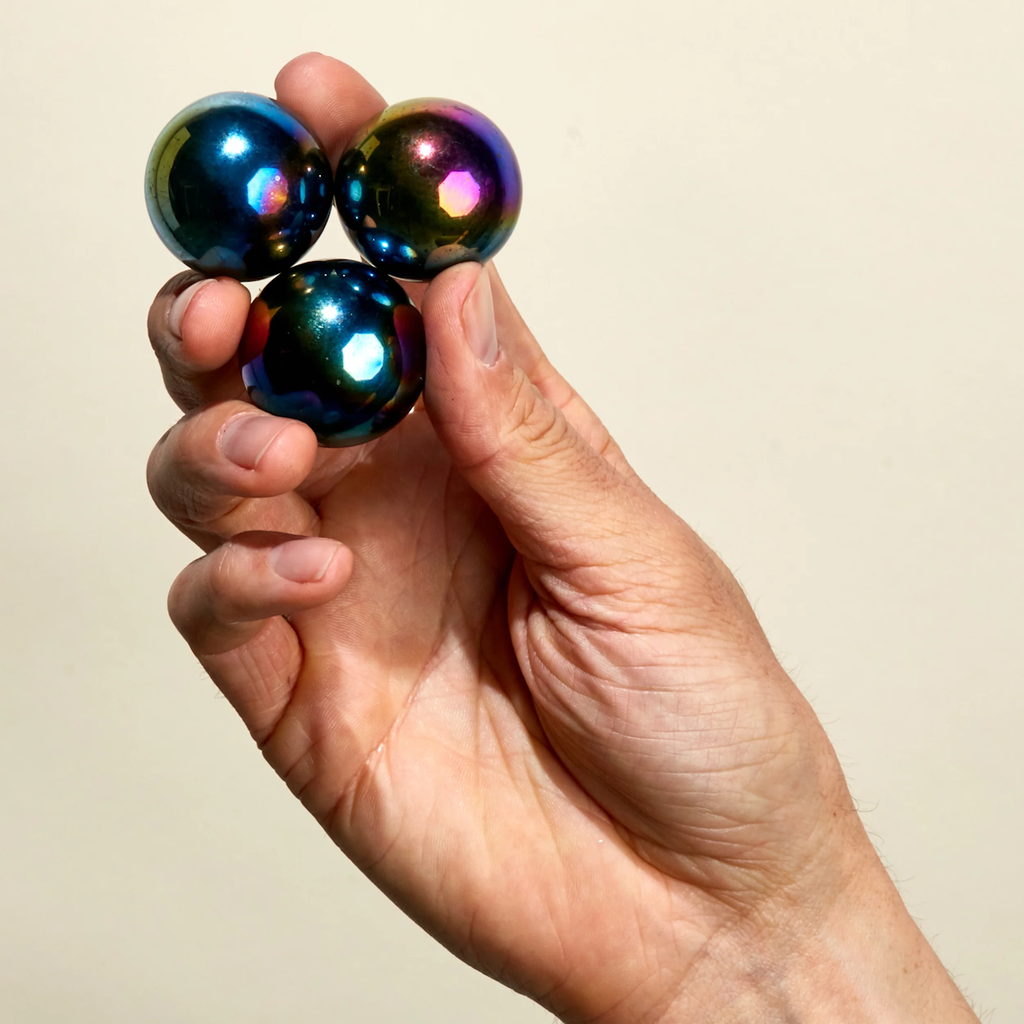 A hand holding three iridescent, metallic spheres with a rainbow sheen against a plain background, conveying a sense of balance and curiosity.