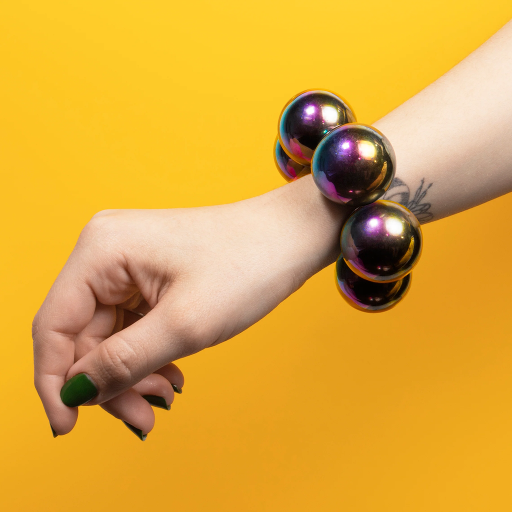 A hand with green painted nails wears a bracelet of large, shiny, multicolored spheres, set against a bright yellow background.