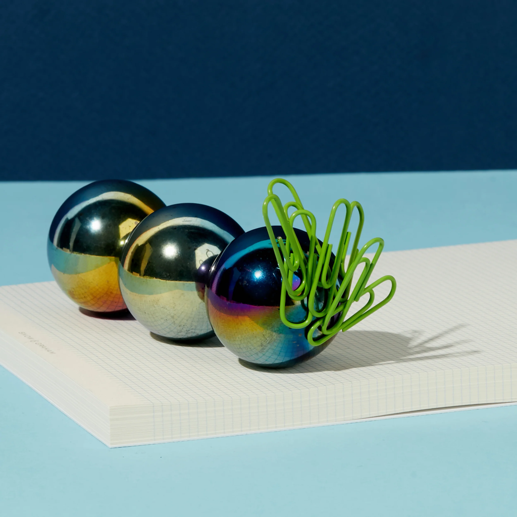 Three shiny metallic spheres in gradient hues sit on graph paper. One sphere has green paper clips attached, casting shadows on a blue background.