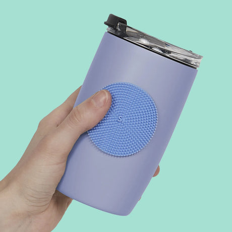 A hand holds a lavender tumbler with a circular light blue dot-textured silicone sensory sticker attached to it.