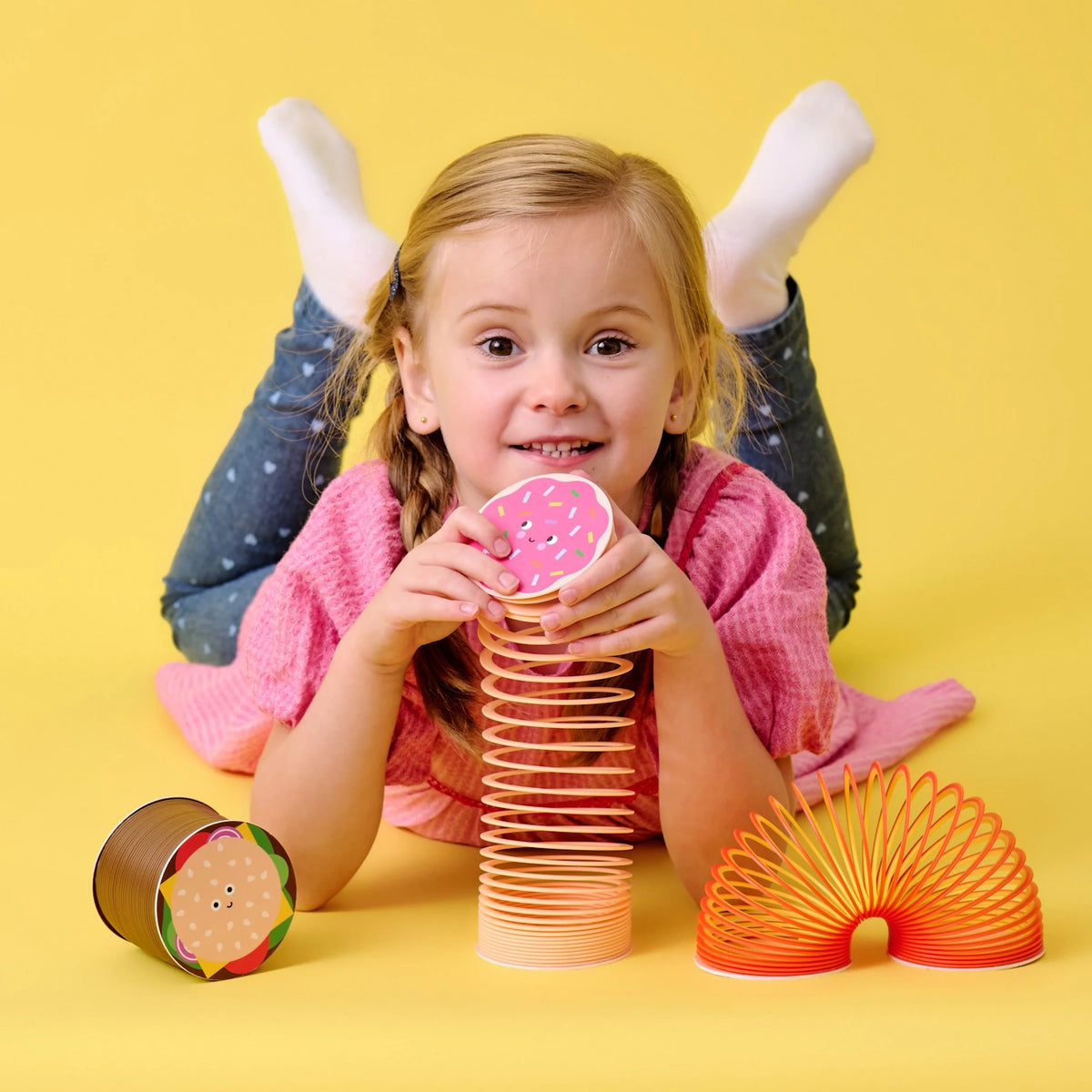A joyful girl with braided hair and a pink sweater lies on the floor, holding a pink donut-shaped toy. Two colorful, coiled toys are beside her. Bright yellow background.