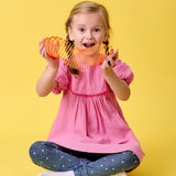 A joyful child in a pink dress sits cross-legged against a yellow background, excitedly playing with a colorful Slinky toy, conveying happiness and playfulness.