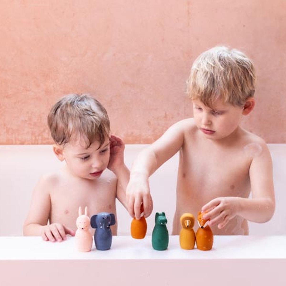 Two young children in a bathtub play with colorful animal-shaped toys. The setting has a warm, playful tone with a soft pink background.