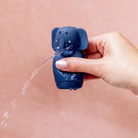 A hand squeezes a blue elephant-shaped toy that squirts water from its trunk, set against a soft pink background. The scene is playful and lighthearted.