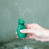 A hand holds a green hippo-shaped bath toy spraying multiple streams of water from its top, set against a blurred gray background, conveying a playful tone.