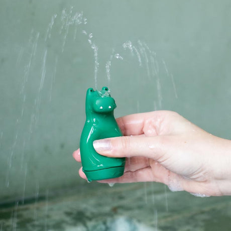 A hand holds a green hippo-shaped bath toy spraying multiple streams of water from its top, set against a blurred gray background, conveying a playful tone.