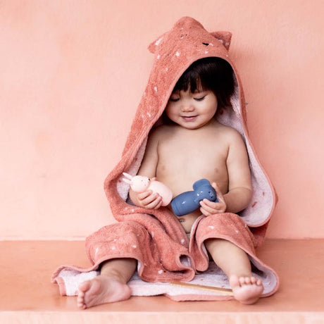 A smiling toddler sits wrapped in a soft, fox-themed hooded towel, holding pink and blue toys. The warm peach backdrop adds a cozy, playful vibe.