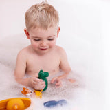 A young child with blonde hair sits in a bubble-filled bath, smiling and playing with colorful bath toys, including a green dinosaur and a yellow boat.