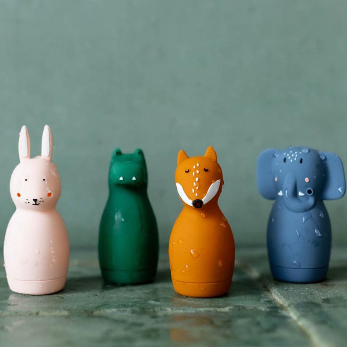 Four colorful animal-shaped soap dispensers stand on a green tile surface: a pink rabbit, green bear, orange fox, and blue elephant, with water droplets.