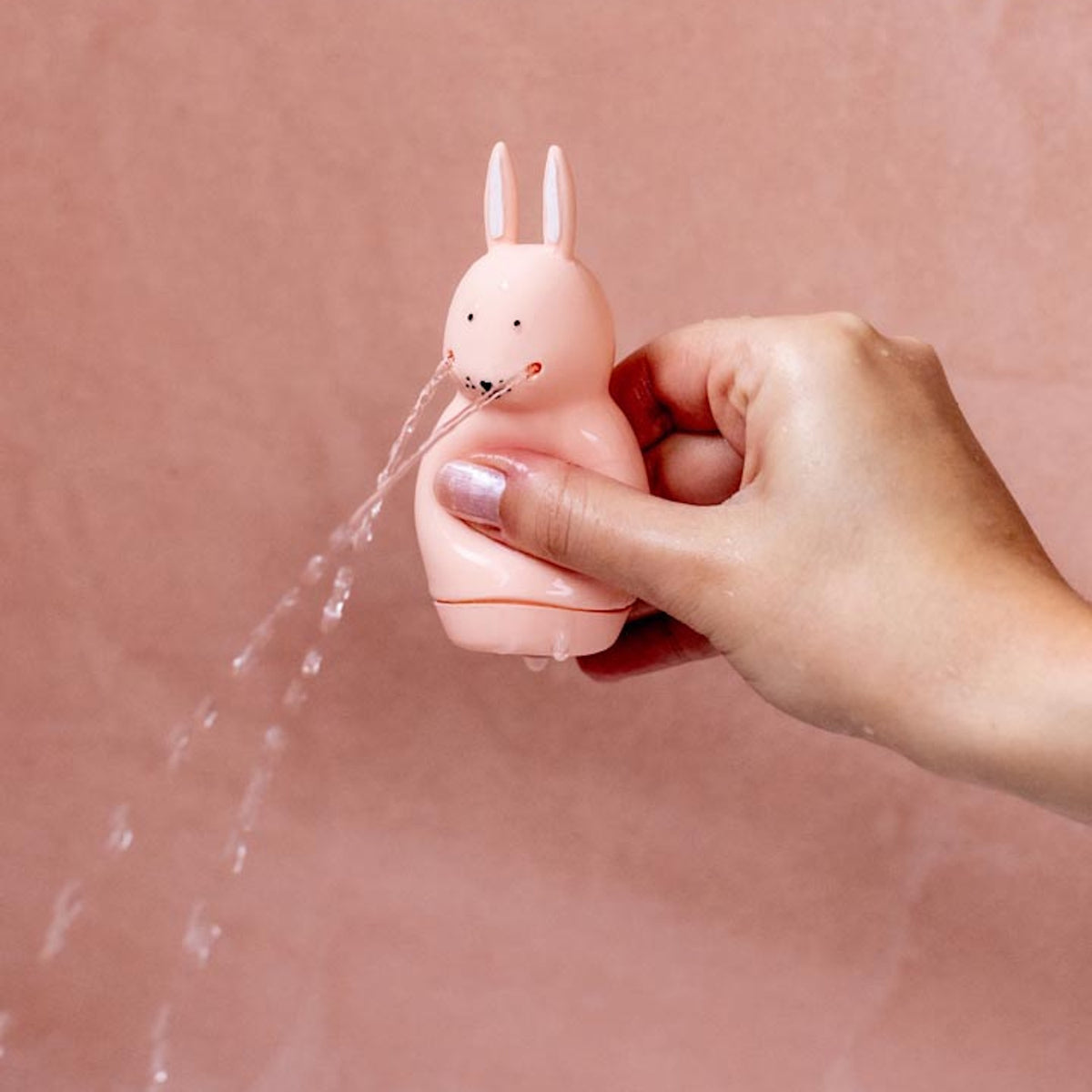 A hand holding a pink rabbit-shaped toy squirting water from its mouth against a soft pink background, creating a playful and whimsical scene.