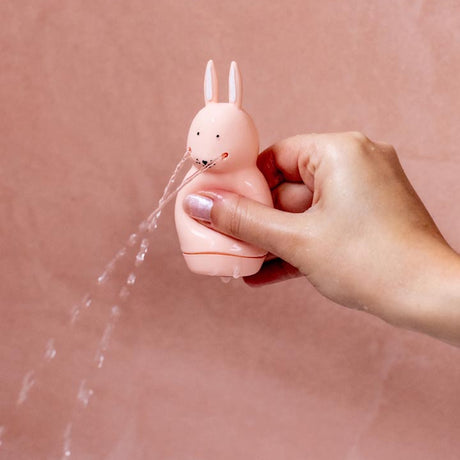 A hand holding a pink rabbit-shaped toy squirting water from its mouth against a soft pink background, creating a playful and whimsical scene.