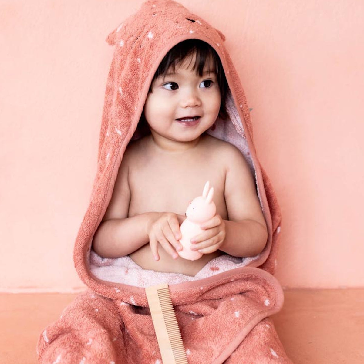 Toddler wrapped in a pink hooded towel with bear ears, holding a pink toy rabbit and a comb, sits against a matching pink background, smiling cheerfully.