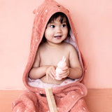Toddler wrapped in a pink hooded towel with bear ears, holding a pink toy rabbit and a comb, sits against a matching pink background, smiling cheerfully.