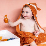 A smiling toddler wrapped in an orange, fox-themed towel plays with a toy in a light room. The background is a soft peach color, creating a warm, cozy mood.