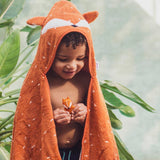 A smiling child in a fox-themed orange towel holds a small toy, surrounded by lush green plants. The scene is playful and warm.