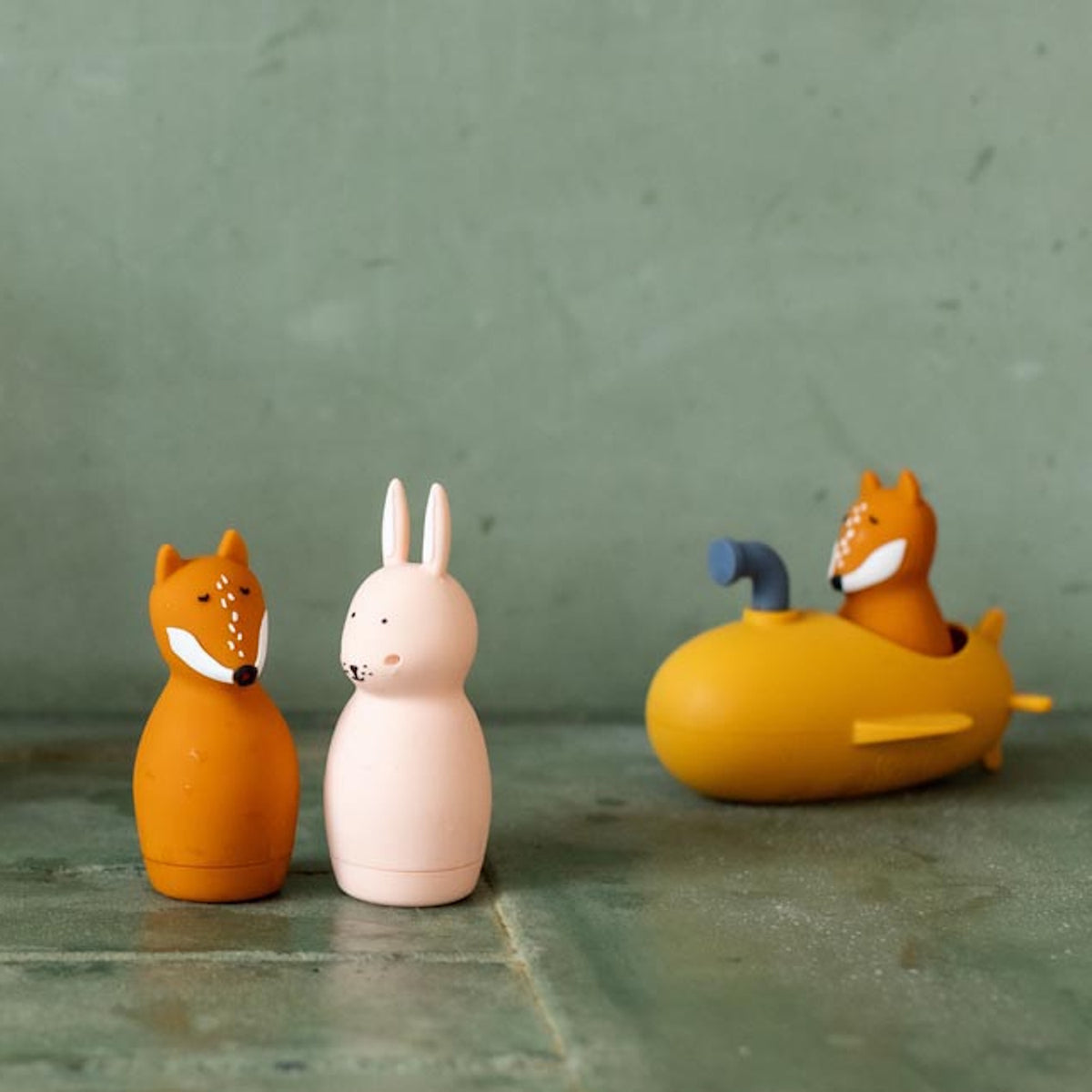 Toy figures of a fox and a rabbit stand on a green surface, with another fox in a yellow submarine toy in the background. The scene is playful and whimsical.