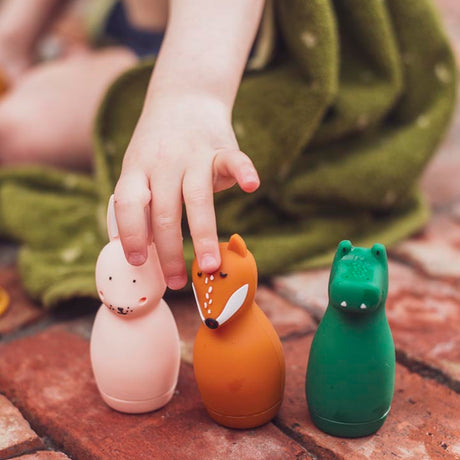 A child's hand reaches for a set of three colorful animal figures on a brick surface: a pink rabbit, an orange fox, and a green crocodile, with a playful, cozy setting.