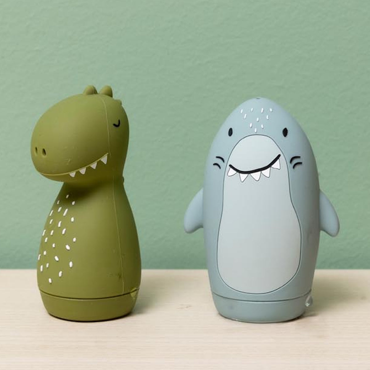 Two cute, egg-shaped rubber toys, a green dinosaur and a blue shark, standing on a light wood surface.