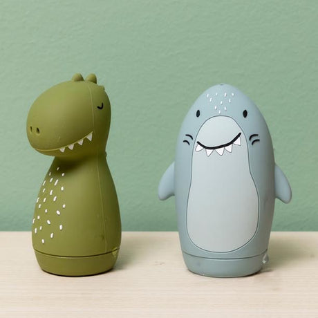 Two cute, egg-shaped rubber toys, a green dinosaur and a blue shark, standing on a light wood surface.