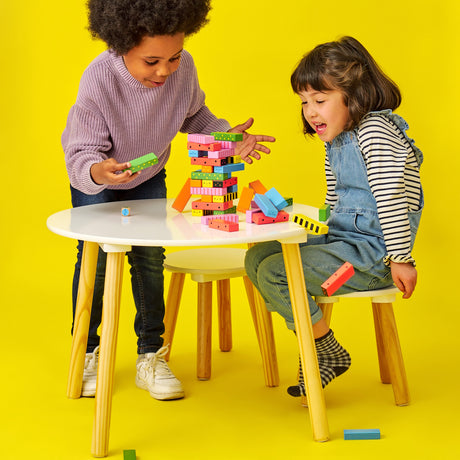 Two children playing a wooden stacking game at a white table, watching colorful blocks fall over against yellow.