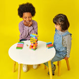 Two happy children sitting at a white table with a colorful wooden block stacking game against a yellow background.