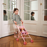 Young girl pushing a toy doll in a pink foldable stroller on a wooden floor in a classic white room.