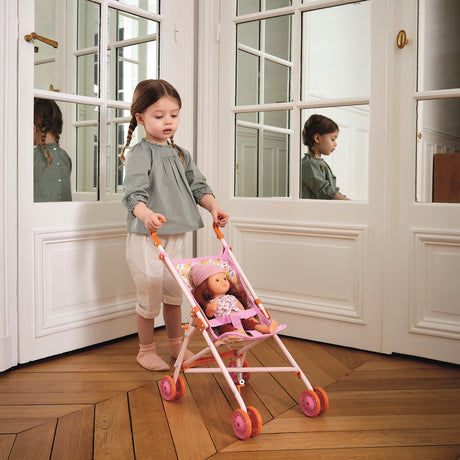 Young girl pushing a toy doll in a pink foldable stroller on a wooden floor in a classic white room.
