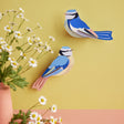 Paper art birds with blue, white, and peach hues on a yellow wall, next to daisies in a pink vase. The scene feels cheerful and artistic.
