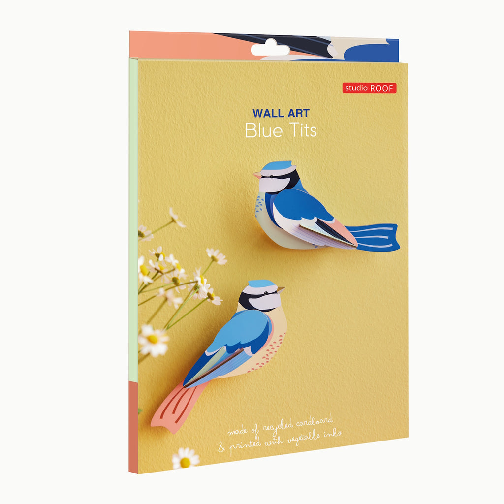 Yellow background showcasing two blue tit birds crafted from layered, colorful cardboard. Text reads "Wall Art Blue Tits" and "made of recycled cardboard & printed with vegetable inks." Flowers accent the bottom left corner.