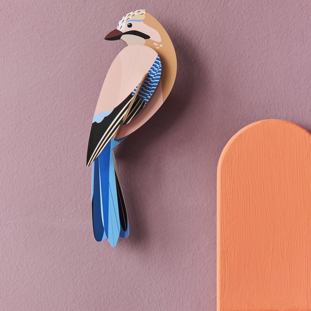A paper-crafted bird with beige, blue, and black feathers is mounted on a light purple wall, next to a semicircular orange shape. The tone is artistic and serene.