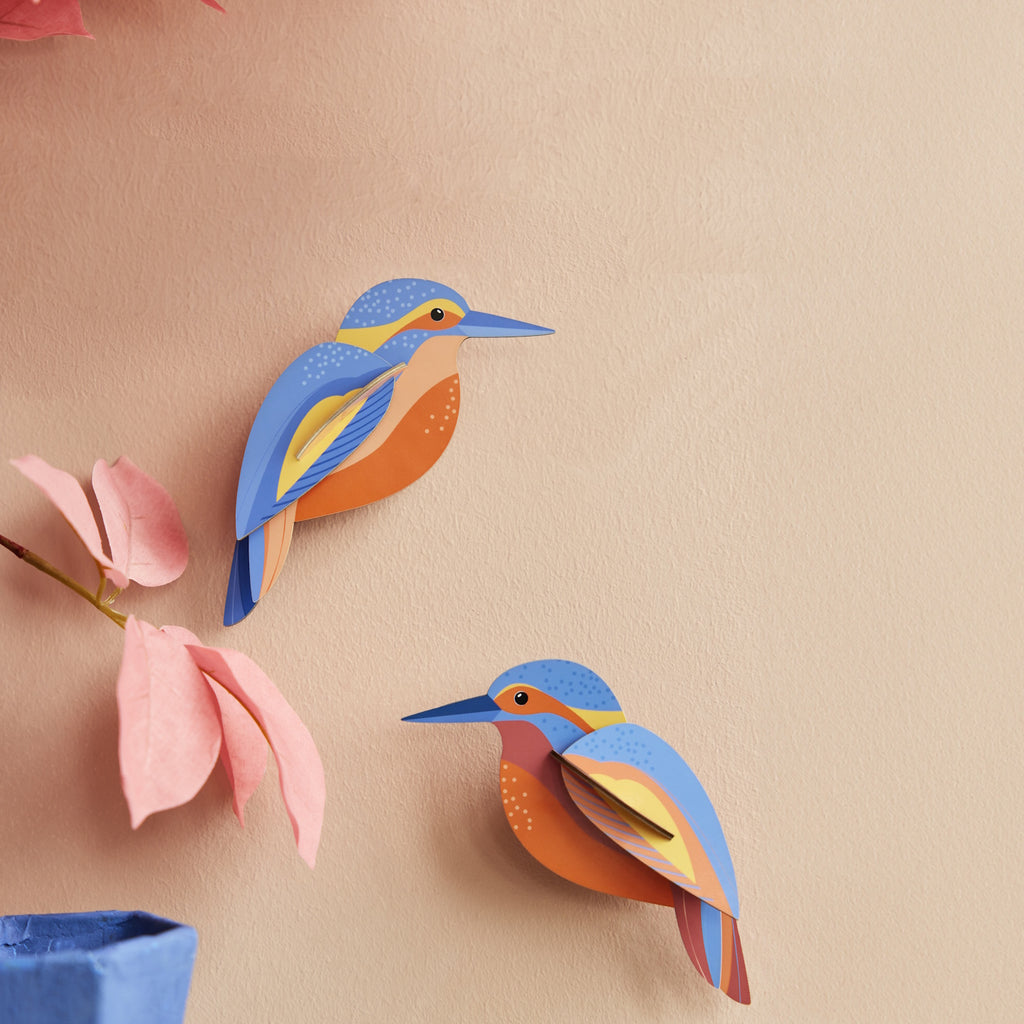 Two colorful paper kingfisher sculptures adorn a beige wall, surrounded by pink paper flowers on the left. The scene feels artistic and serene.
