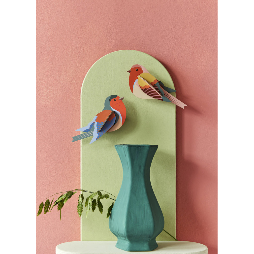Colorful paper birds are mounted on a pastel green panel against a pink wall. A teal vase with ferns is in the foreground, creating a vibrant and artistic display.
