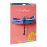 A vibrant blue dragonfly wall decoration on a coral background with yellow text and a small studio logo in red. The dragonfly is made of layered recycled cardboard, showing intricate wing patterns and a segmented body.