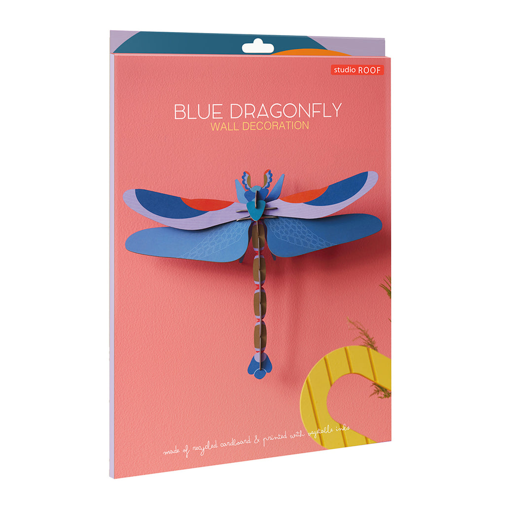 A vibrant blue dragonfly wall decoration on a coral background with yellow text and a small studio logo in red. The dragonfly is made of layered recycled cardboard, showing intricate wing patterns and a segmented body.