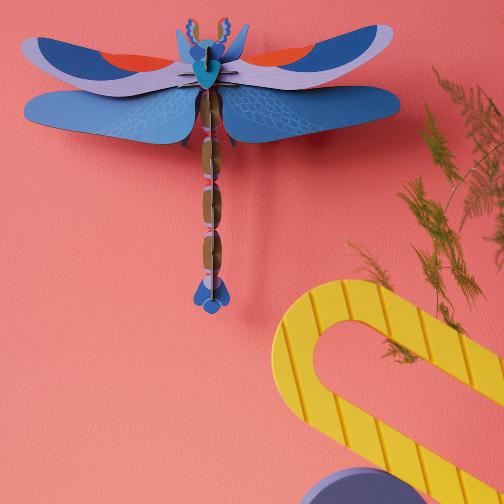 Papercraft dragonfly with vibrant blue, purple, and orange wings on a pink wall. Nearby, a yellow graphic and green foliage add dynamic contrast.