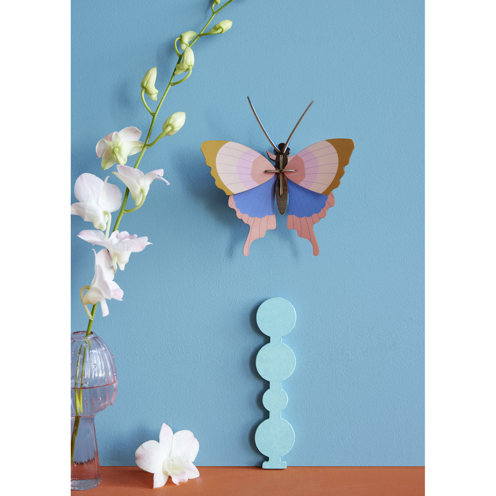 A colorful paper butterfly and white orchids are set against a light blue wall. A decorative blue object and glass vase add elegance to the serene scene.