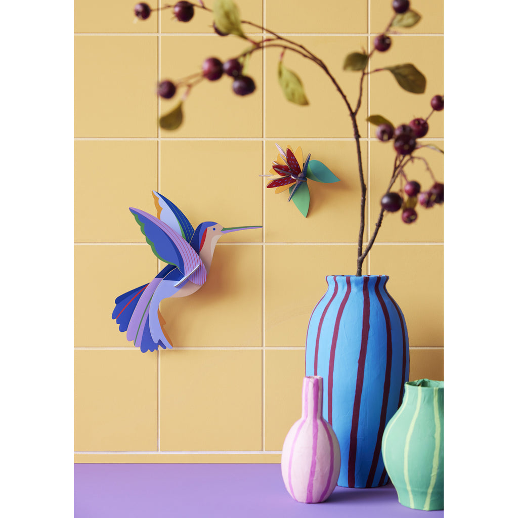 Vibrant paper hummingbird and flower on a yellow tiled wall, with vases in blue, pink, green and a twig with berries, creating a lively, artistic scene.