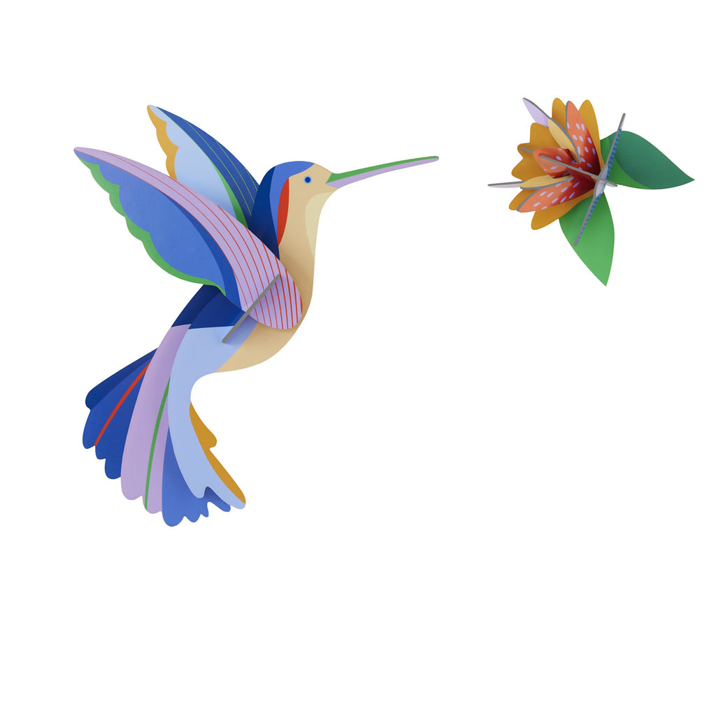 A colorful, stylized paper hummingbird with vibrant blue, pink, and yellow wings approaches an intricately crafted orange and green flower. Bright and whimsical.