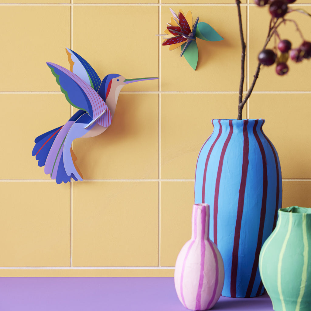 Vibrant paper hummingbird and flower on a yellow tiled wall, with vases in blue, pink, green and a twig with berries, creating a lively, artistic scene.