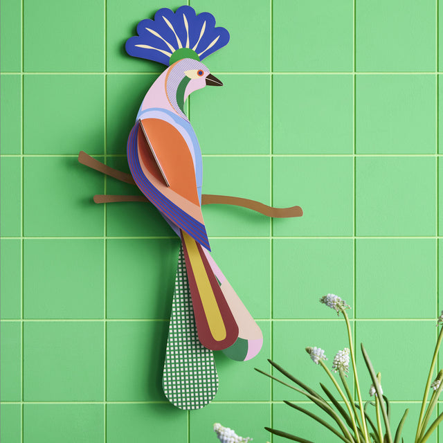 A colorful, stylized bird art sculpture on a light green tiled wall. The bird is perched on a branch, with a vibrant plumage and geometric patterns.